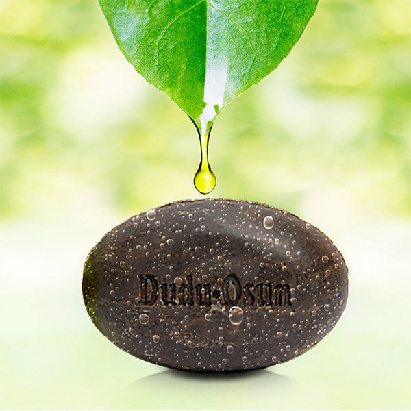 A dark brown polished stone engraved with 'Dudu Osun' sits on a reflective surface, with a single green leaf hanging above and a clear oil drop dripping onto the stone against a soft green blurred background.