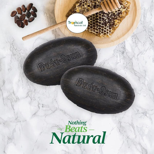 Two oval black soap bars engraved with 'Dudu-Osun' on a white marble surface, with a wooden honeycomb slice, honey dipper, and cocoa beans in the background. A circular 'Tropical Naturals Ltd' logo appears near the top, and 'Nothing Beats Natural' is written at the bottom.